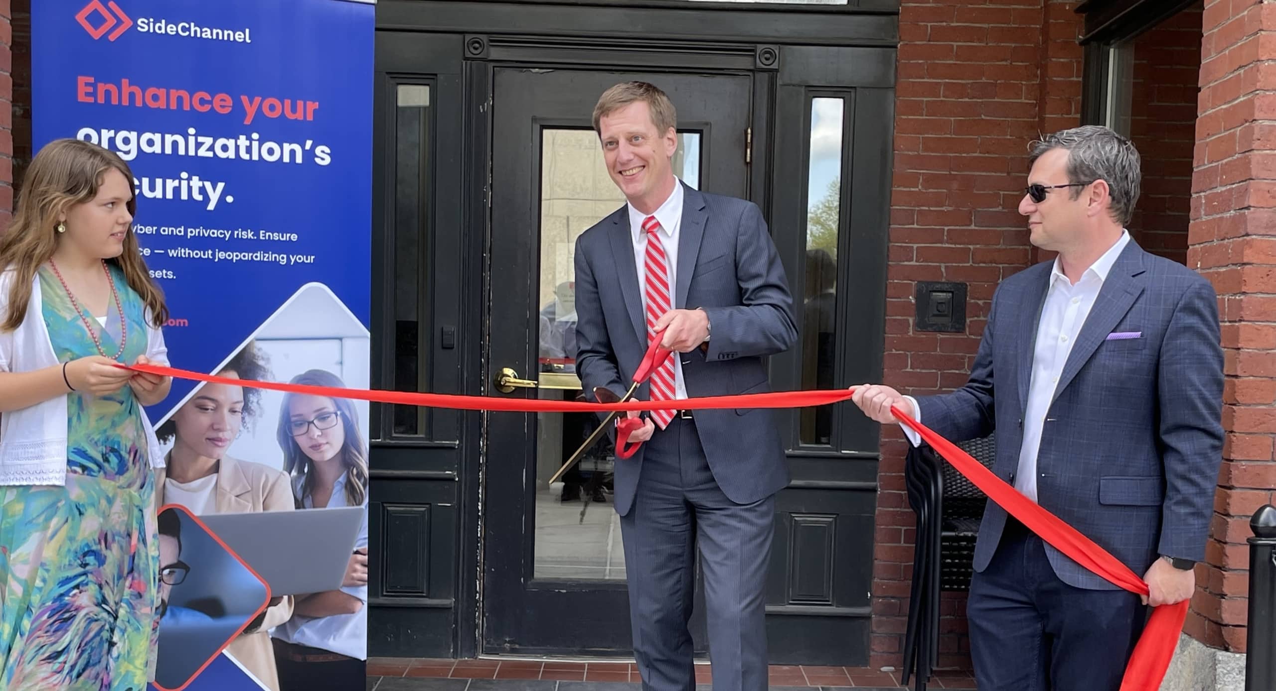a man in a blue suit and a young girl in a green dress hold a red ribbon across a doorway and third person holds a pair of very large scissors and prepares to cut the red ribbon in half