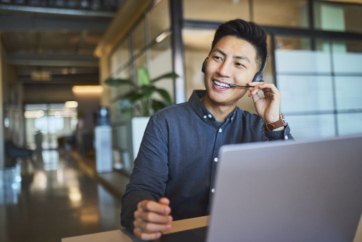 Happy Asian customer service agent wearing a headset and working on a laptop in a modern office environment.