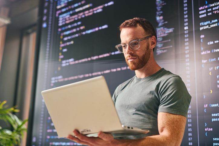 Software developer working on a laptop with code displayed on a large screen in a modern office.