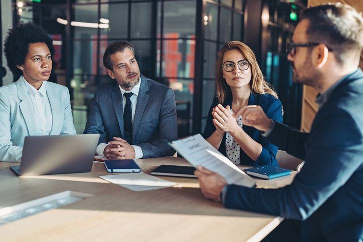 Four professionals sit around a table in a modern office, engaged in a serious discussion with documents and a laptop.