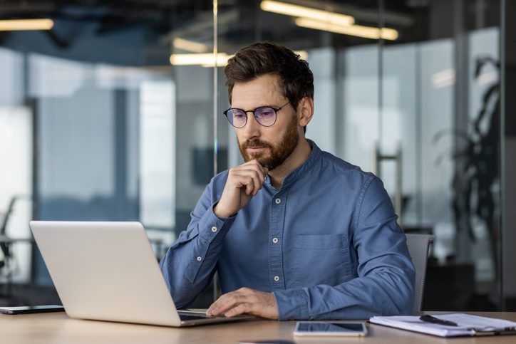 A man in glasses and a blue shirt sits at a desk, thoughtfully looking at a laptop in a modern office setting.