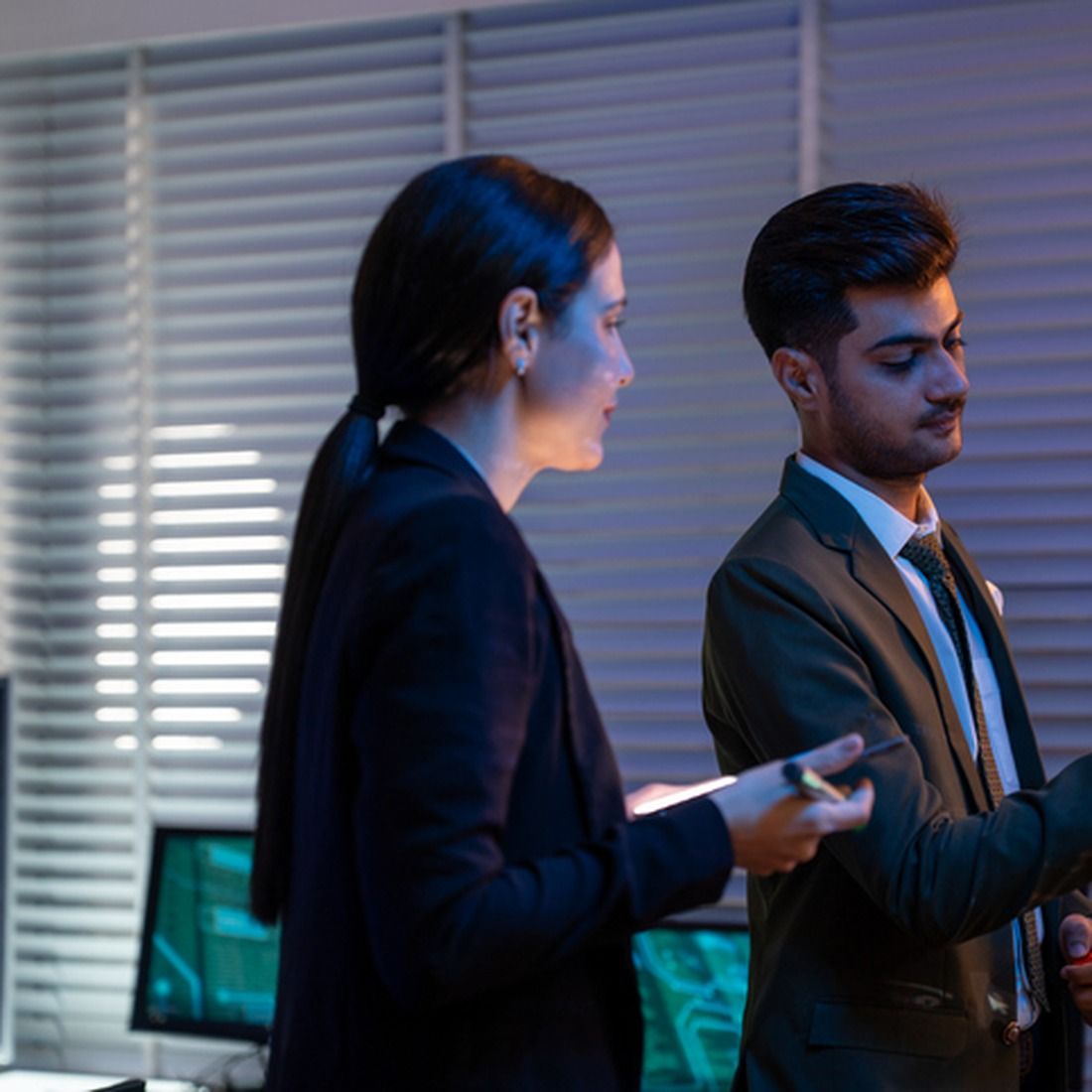 Two professionals in business attire stand side by side, looking at their devices in a modern office setting.