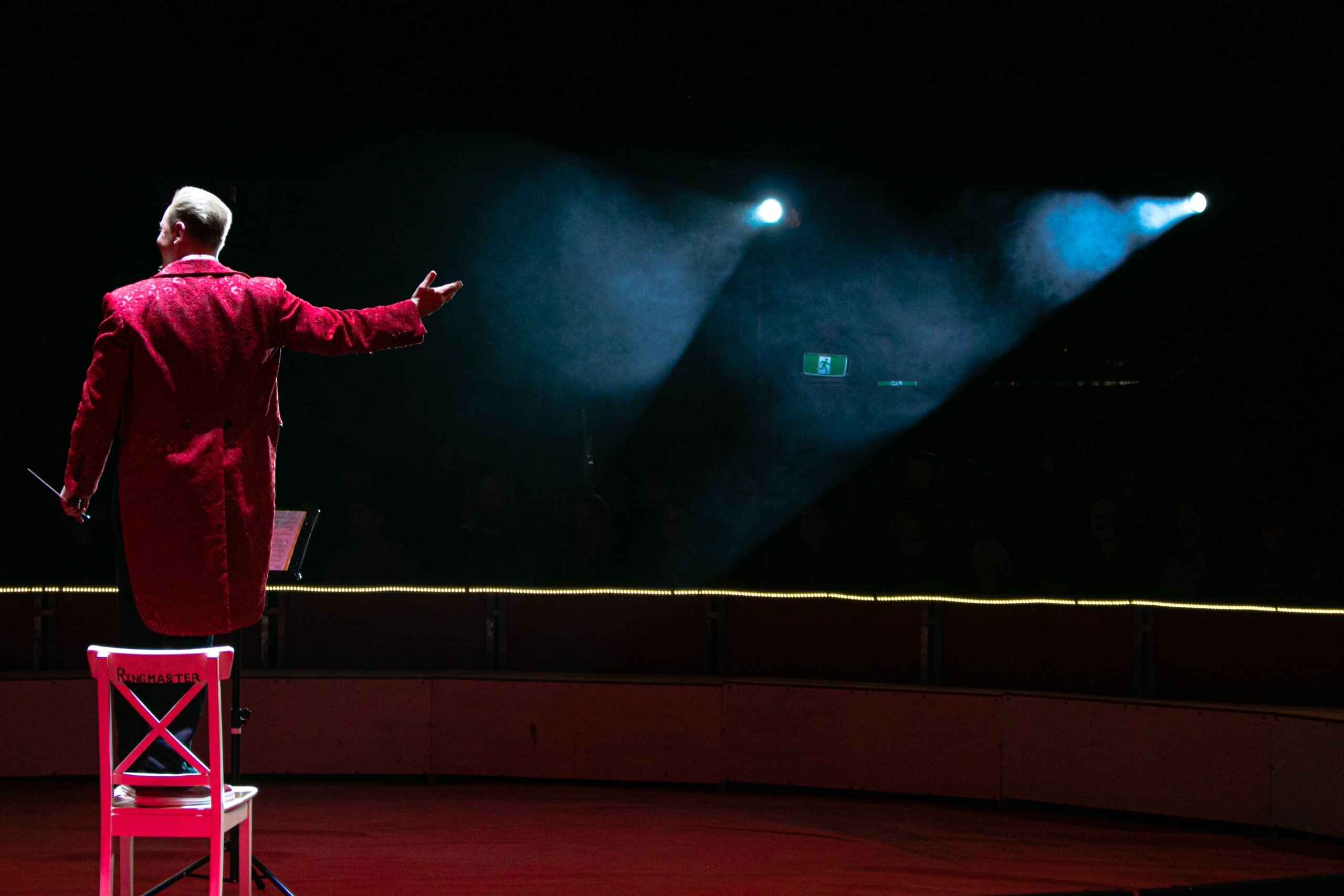 man in a red tuxedo jacket with tails conducting an orchestra we cannot see while standing on a red chair in front of stage lights in the background